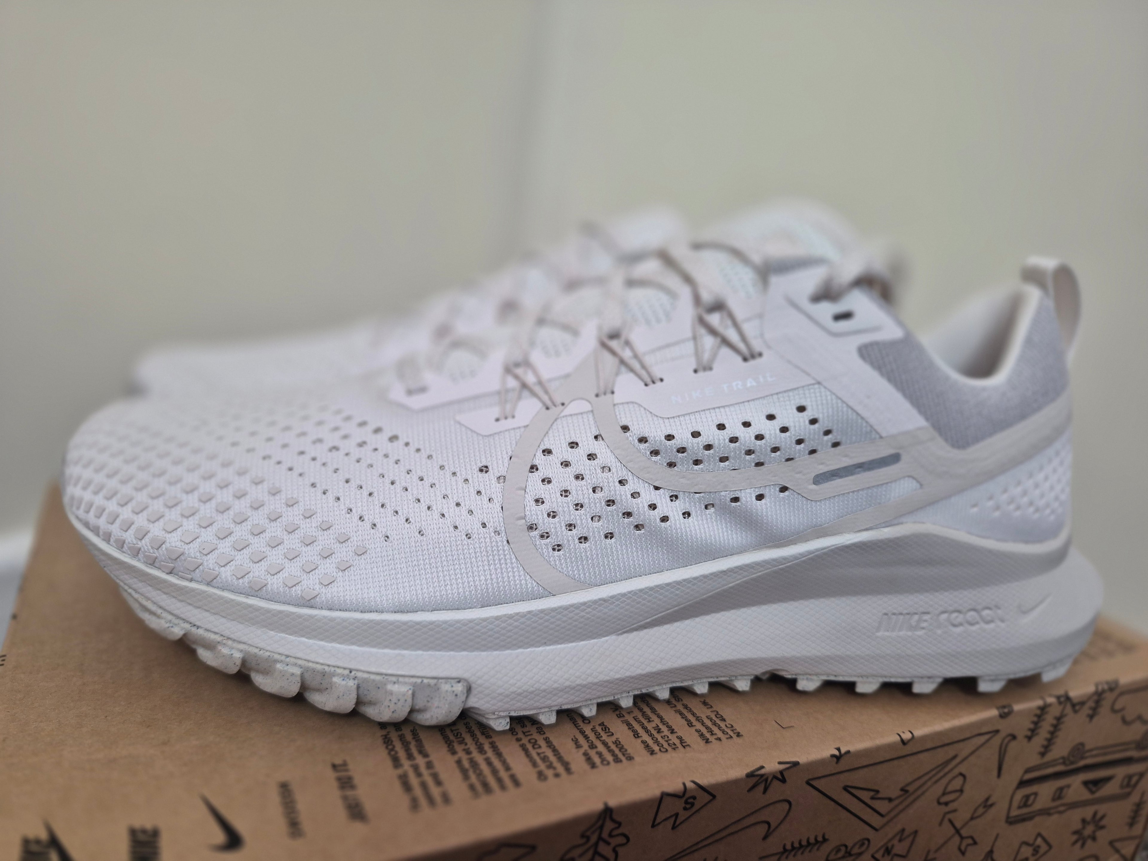 A pair of white Nike running shoes with perforated mesh uppers and thick cushioned soles, displayed on top of a brown shoebox.