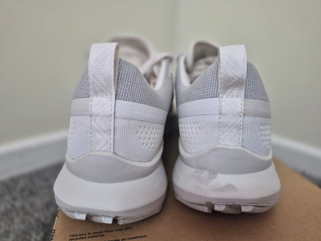 A pair of white and light gray athletic sneakers shown from the back, resting on a cardboard box against an indoor wall and floor background.