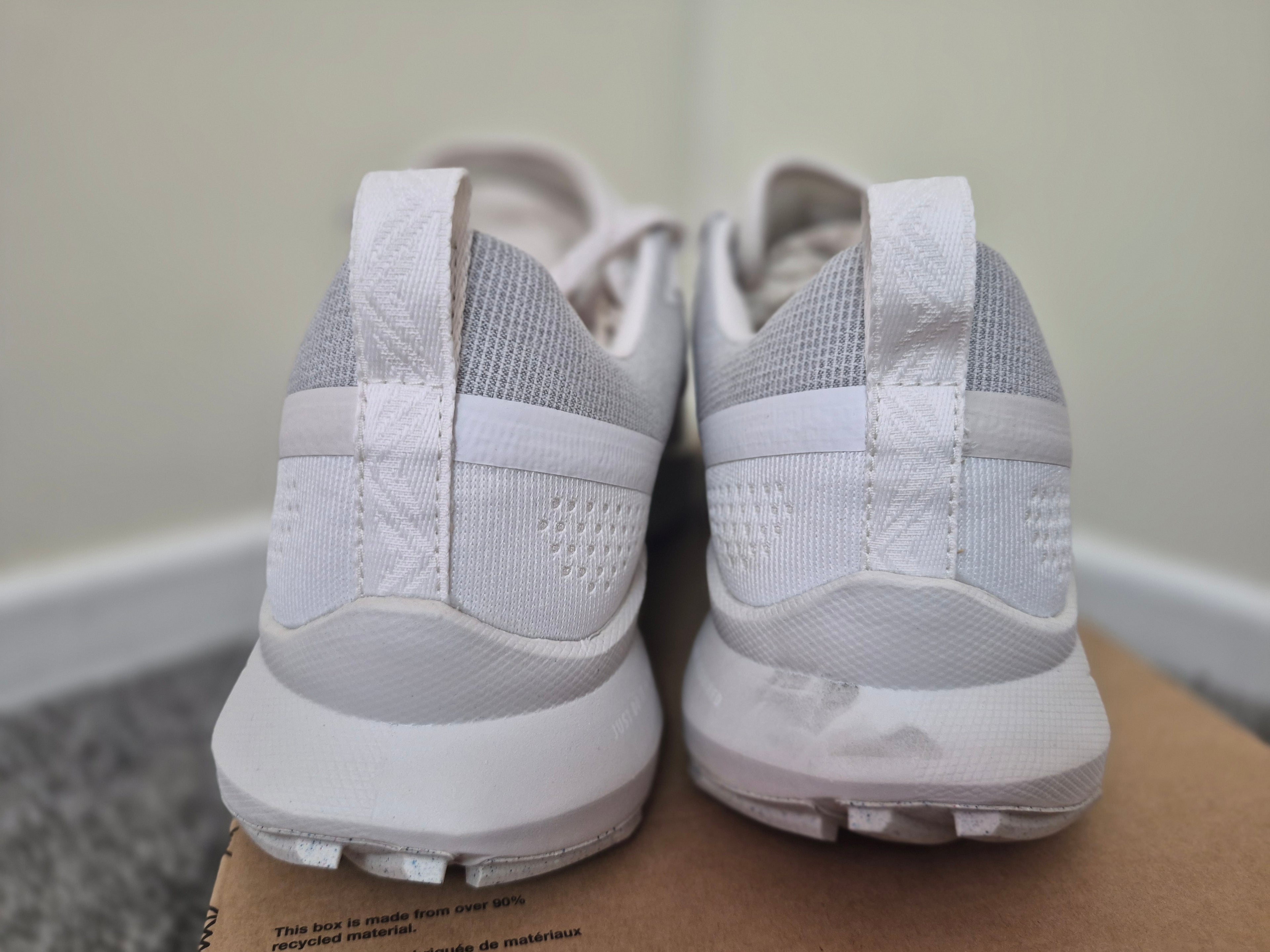 A pair of white and light gray athletic sneakers shown from the back, resting on a cardboard box against an indoor wall and floor background.